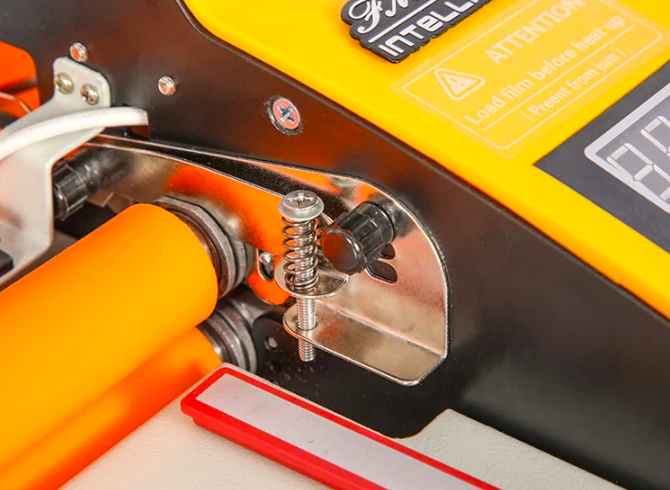 Détail du mécanisme de ressort et des rouleaux de la plastifieuse FM-3810 avec étiquette de sécurité.